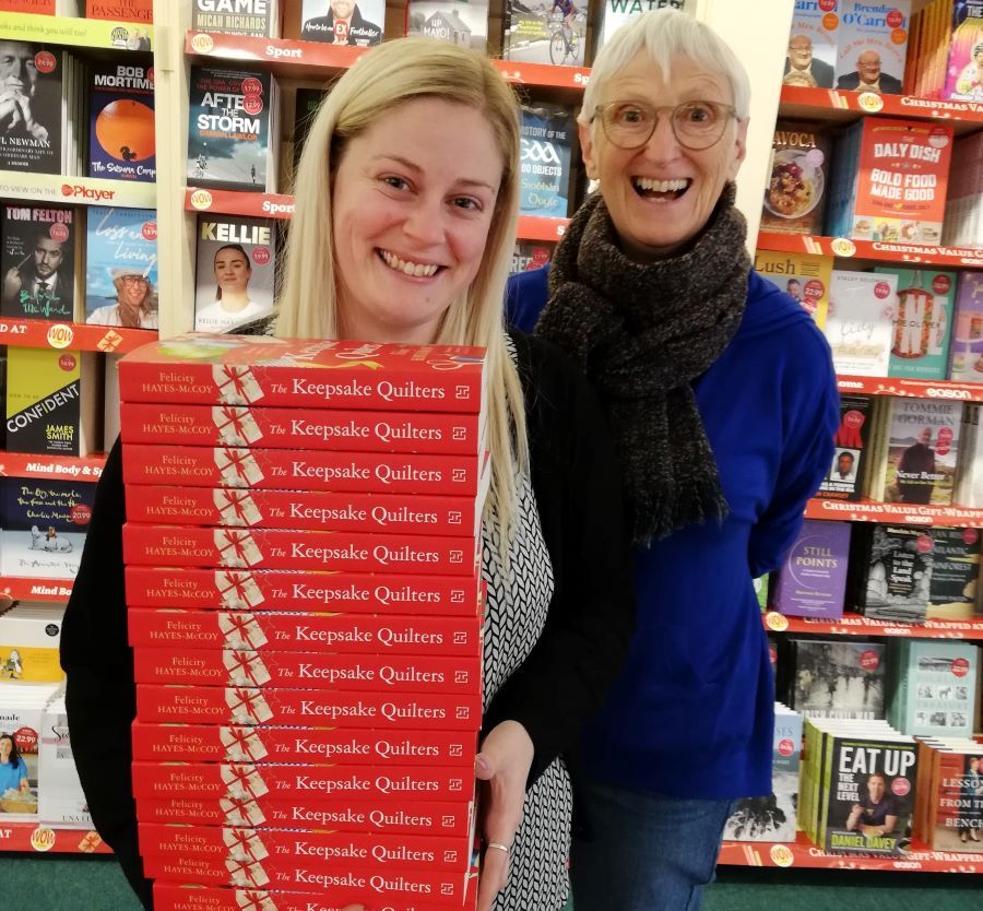 Image of a young, fair-haired bookseller holding a pile of copies of author Felicity Hayes-McCoy's 2022 novel THE KEEPSAKE QUILTERS, with the author behind her and bookshop shelves in the background