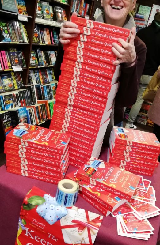 A stack of copies of author Felicity Hayes-McCoy's 2022 novel THE KEEPSAKE QUILTERS against a bookshop background